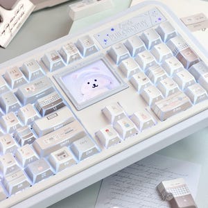 May include: A white mechanical keyboard featuring custom keycaps with various icons and patterns. A central square display screen shows a cartoon bear character. The top panel includes the text Love's PHILOSOPHY with star accents.