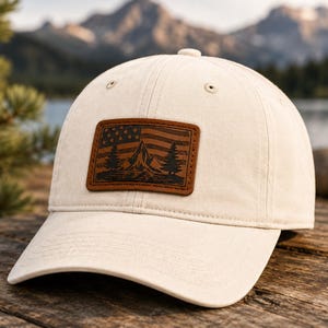 Könnte beinhalten: Eine cremefarbene Baseballkappe mit einem rechteckigen braunen Lederaufnäher auf der Vorderseite. Der Aufnäher ist mit einem grafischen Design geprägt, das ein Gebirge, Tannenbäume und ein stilisiertes amerikanisches Flaggendesign zeigt.