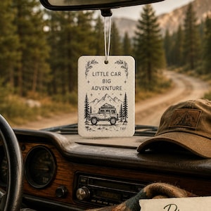 May include: A rectangular white air freshener hanging from a rearview mirror, featuring a black line art illustration of a car, mountains, and pine trees with the text LITTLE CAR BIG ADVENTURE printed in the centre and the letters TRF at the bottom.