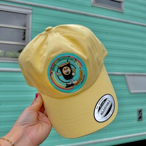 May include: A light yellow baseball cap featuring a circular patch with a cartoon coffee cup character and the text Stay Caffeinated and Groovy Brew. The cap includes a black and white sticker on the brim that reads The Authentic Adjustable.