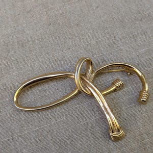 May include: A gold-tone metal brooch featuring a continuous, looping design that forms an abstract, elongated shape. The piece has a polished, reflective surface with textured, ribbed detailing at the two rounded ends of the metal structure.