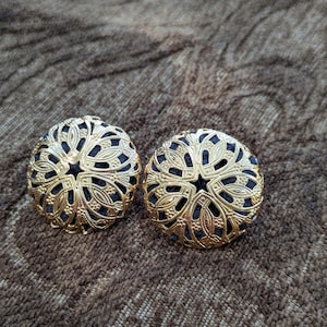 May include: A pair of round, gold-tone metal buttons featuring an intricate, openwork filigree design. The metallic overlay displays a detailed floral-inspired pattern with cutouts that reveal a dark, solid-colored fabric base underneath.
