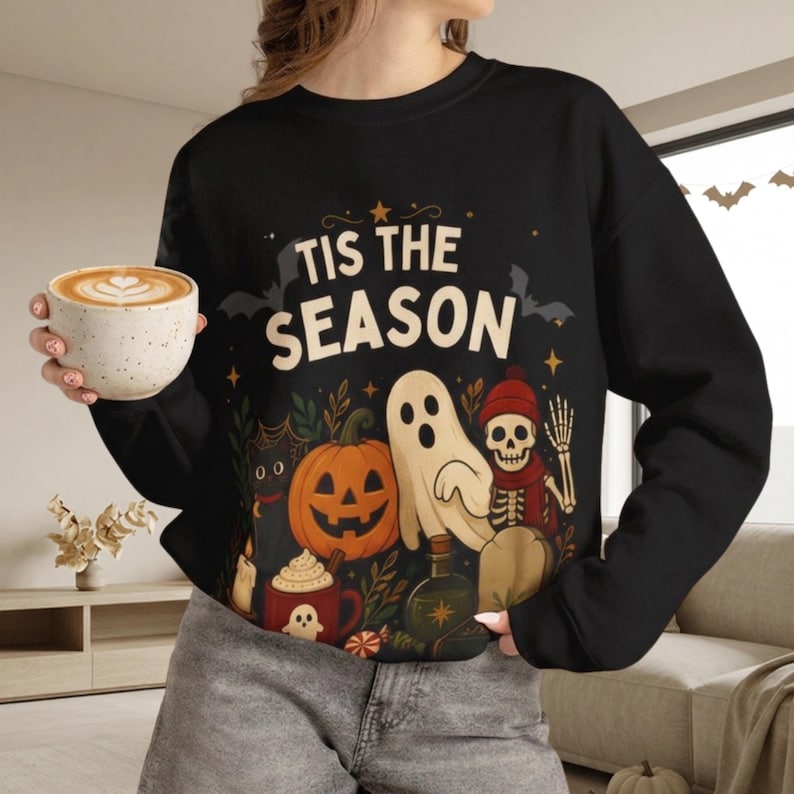 May include: A black long-sleeve sweatshirt featuring the text 'TIS THE SEASON' in cream letters. The graphic includes a jack-o'-lantern, a white ghost, a skeleton wearing a red hat and scarf, a black cat, a red mug, a candle, and bats.