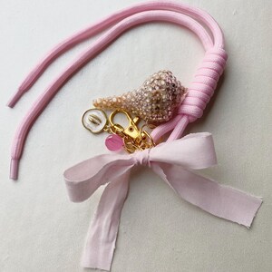 Könnte beinhalten: Ein hellrosa Kordelband mit einem goldfarbenen Karabinerverschluss, einer mit kleinen klaren Kristallen besetzten Muschel, einem kleinen weißen runden Anhänger mit goldenem Muschelsymbol, einer transparenten rosa Perle und einer hellrosa Stoffschleife.