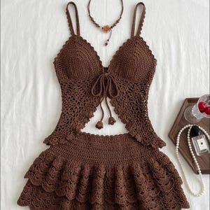May include: A dark brown crochet two-piece set consisting of a sleeveless top with thin straps, a front tie closure, and a matching tiered mini skirt with a scalloped hem. A thin beaded necklace with a small flower charm is positioned above the top.