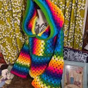 May include: A hooded sweater crafted from multicolored yarn in a granny square crochet pattern. The garment features a vibrant spectrum of red, orange, yellow, green, blue, and purple hues arranged in repeating blocks, with a large hood and long sleeves.