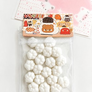 May include: A clear plastic bag filled with small, white, paw-shaped items. The top of the bag features a cardboard header with a brown bear character, sushi, and octopus illustrations, along with Japanese text including "いただきます" and "おにぎり".
