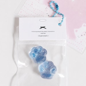 May include: Two translucent blue paw-shaped charms inside a clear plastic bag with a white header card. The card features a small black bow icon and the text: If I'm in the mood for the weather, I'll be stuck in a cloudy day. ( Sweet dreams ).