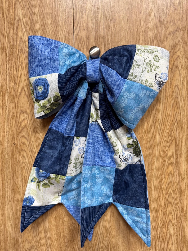 Pu&ograve; includere: Un grande fiocco decorativo realizzato con toppe di tessuto trapuntato in varie tonalit&agrave; di blu, bianco e verde. Il tessuto presenta motivi di rose, farfalle e fogliame. Un piccolo bottone metallico &egrave; centrato nella parte superiore del fiocco.