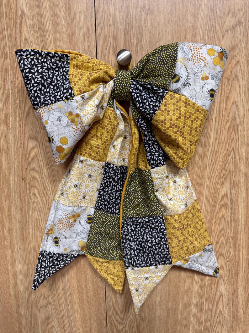 May include: A large decorative bow made from quilted fabric patches featuring a bee and honeycomb theme. The fabric displays a mix of black, white, and yellow patterns, including floral motifs, geometric honeycombs, and small bee illustrations.