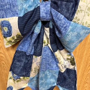 Pu&ograve; includere: Un grande fiocco in tessuto realizzato con vari pezzi trapuntati in tonalit&agrave; di blu, bianco e verde. Il design patchwork presenta motivi floreali, stampe botaniche e un motivo a farfalla. Il fiocco &egrave; fissato da un nodo centrale in tessuto.