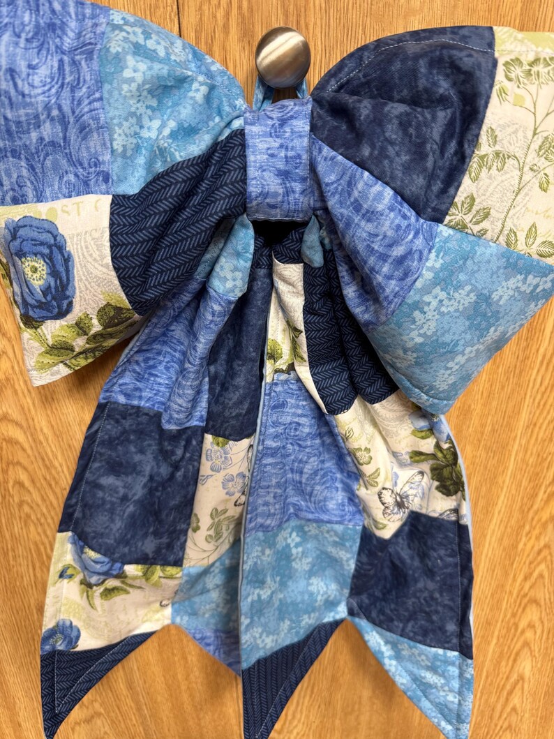 Pu&ograve; includere: Un grande fiocco in tessuto realizzato con vari pezzi trapuntati in tonalit&agrave; di blu, bianco e verde. Il design patchwork presenta motivi floreali, stampe botaniche e un motivo a farfalla. Il fiocco &egrave; fissato da un nodo centrale in tessuto.