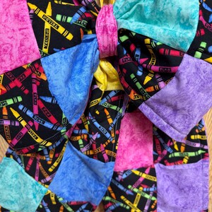 May include: A large fabric hair bow featuring a black background with a colorful crayon print and solid-colored patches in pink, turquoise, blue, and purple. The center is cinched with a pink fabric band, and it hangs from a silver metal hook.
