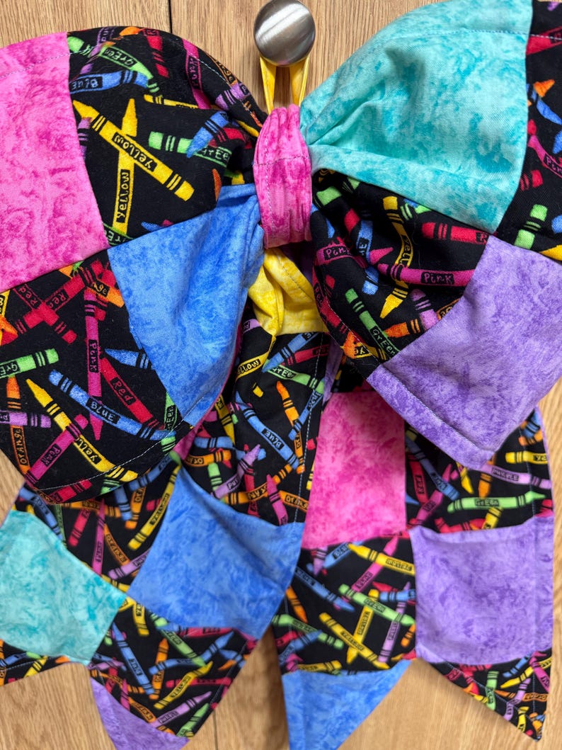 May include: A large fabric hair bow featuring a black background with a colorful crayon print and solid-colored patches in pink, turquoise, blue, and purple. The center is cinched with a pink fabric band, and it hangs from a silver metal hook.