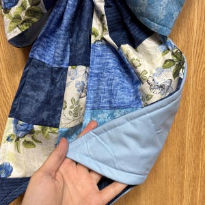 Pu&ograve; includere: Un pezzo di tessuto trapuntato con un design patchwork di vari motivi blu e bianchi, inclusi disegni floreali, farfalle e texture a spina di pesce. Il retro &egrave; in tessuto azzurro tinta unita con cuciture visibili.