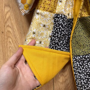 May include: A quilted jacket featuring a patchwork design of various fabrics in yellow, black, and white. The patterns include honeycombs, bees, and leaf motifs. The garment has a solid yellow lining and a visible stitched seam along the edge.