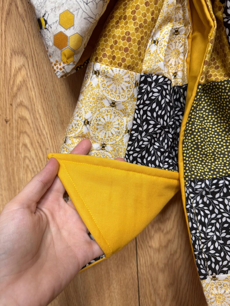 May include: A quilted jacket featuring a patchwork design of various fabrics in yellow, black, and white. The patterns include honeycombs, bees, and leaf motifs. The garment has a solid yellow lining and a visible stitched seam along the edge.
