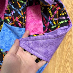 May include: A quilted fabric item featuring a black background with a colorful crayon print, accented by solid patches of mottled purple, blue, and pink fabric. The edges are finished with a purple binding, and the construction shows visible stitching.