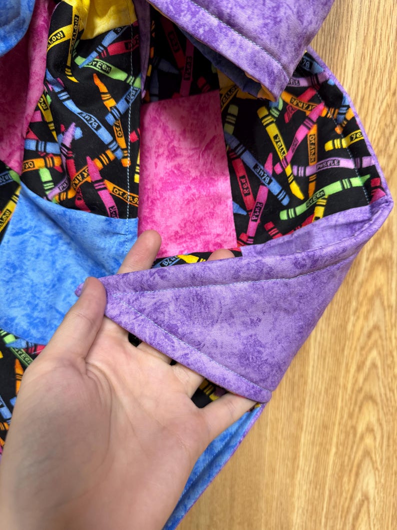 May include: A quilted fabric item featuring a black background with a colorful crayon print, accented by solid patches of mottled purple, blue, and pink fabric. The edges are finished with a purple binding, and the construction shows visible stitching.