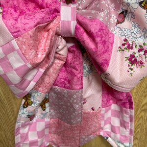 May include: A large fabric bow featuring a patchwork design in various shades of pink and white. The textile includes patterns of checkered squares, floral motifs, and butterflies. The bow is constructed with multiple quilted fabric segments.