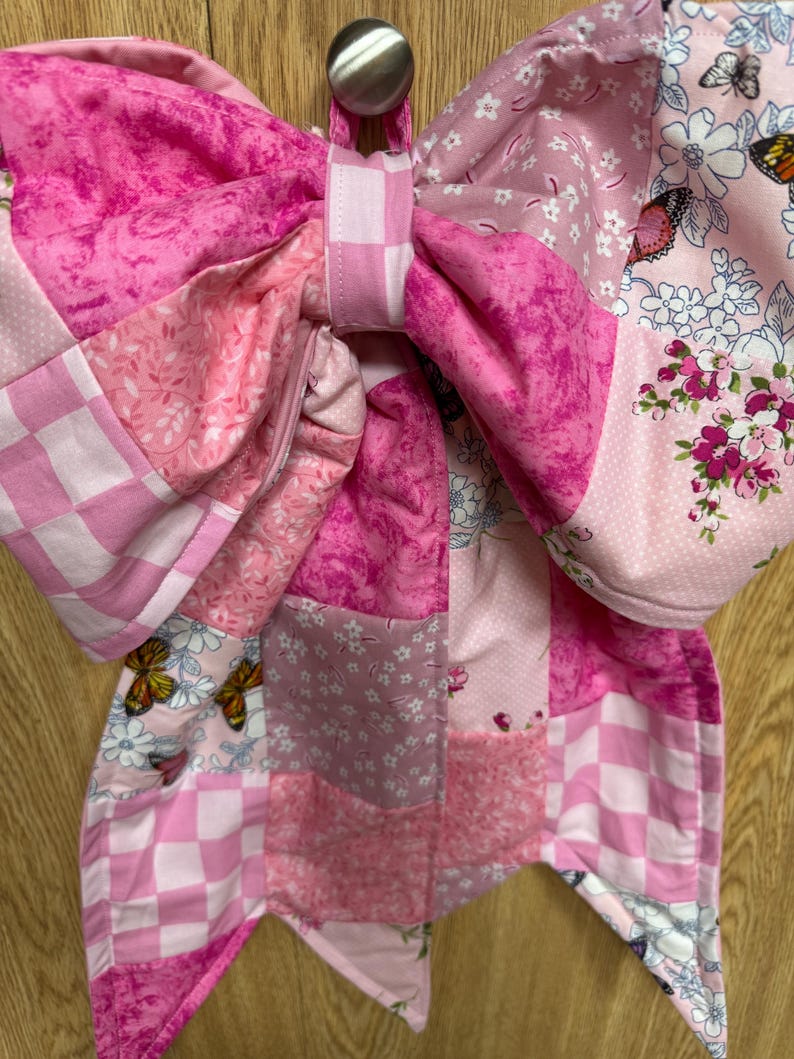 May include: A large fabric bow featuring a patchwork design in various shades of pink and white. The textile includes patterns of checkered squares, floral motifs, and butterflies. The bow is constructed with multiple quilted fabric segments.