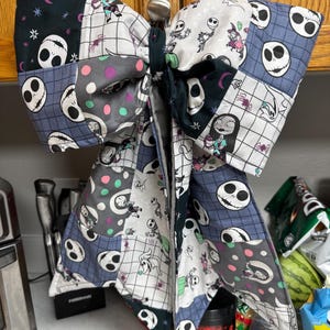 Puede incluir: Un lazo de tela grande con un diseño de patchwork que incluye varios patrones como caras de Jack Skellington, Sally, Zero, arañas y lunares de colores. El lazo está construido con múltiples cuadrados de tela acolchada en negro, blanco y gris.