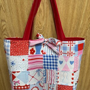 Puede incluir: Un bolso tote de patchwork con una variedad de cuadrados de tela en patrones rojos, blancos y azules, incluyendo lunares, cuadros vichy, rayas y motivos florales. El bolso tiene dos asas largas de tela roja y un lazo decorativo en el centro.
