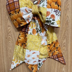 May include: A large decorative bow made from quilted fabric featuring a patchwork design. The pattern includes orange pumpkins, yellow autumn leaves, and brown plaid squares. The bow has two long, pointed tails and a central knot detail.