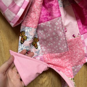 May include: A quilted fabric item featuring a patchwork design in various shades of pink. The pattern includes squares with floral motifs, butterfly illustrations, and a pink and white checkered print, all stitched together with visible seams.