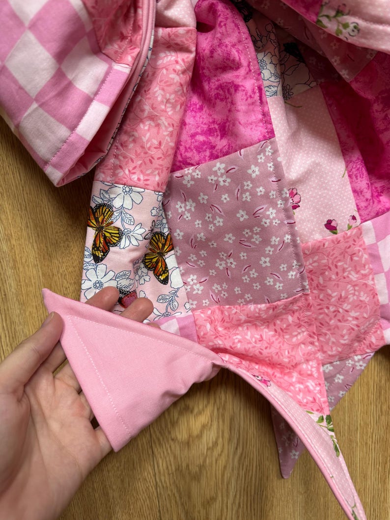 May include: A quilted fabric item featuring a patchwork design in various shades of pink. The pattern includes squares with floral motifs, butterfly illustrations, and a pink and white checkered print, all stitched together with visible seams.