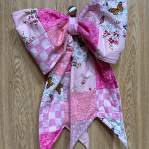 Pode incluir: Um grande laço de tecido com um design de patchwork em vários tons de rosa. Os padrões do tecido incluem quadrados xadrez, estampas florais e motivos de borboletas. O laço é construído com vários painéis de tecido e tem duas pontas.