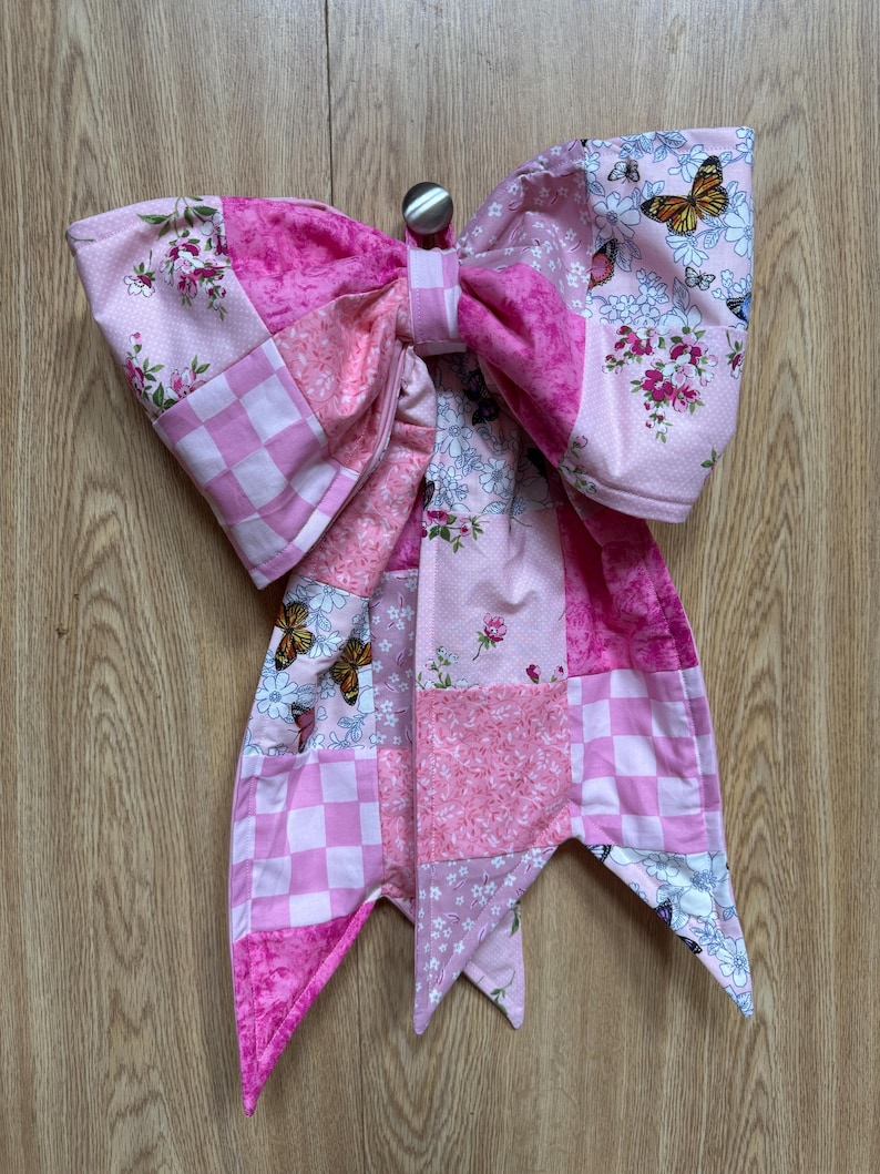 May include: A large fabric bow featuring a patchwork design in various shades of pink. The fabric patterns include checkered squares, floral prints, and butterfly motifs. The bow is constructed with multiple fabric panels and has two long tails.