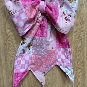 May include: A large decorative bow made from quilted fabric featuring a patchwork design in various shades of pink. The fabric patterns include checkered squares, floral prints, and butterfly motifs. A small silver-colored metal button is at the center.