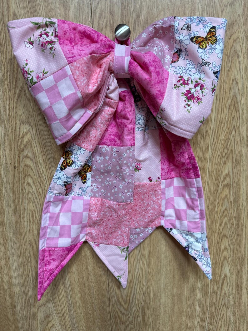 May include: A large decorative bow made from quilted fabric featuring a patchwork design in various shades of pink. The fabric patterns include checkered squares, floral prints, and butterfly motifs. A small silver-colored metal button is at the center.