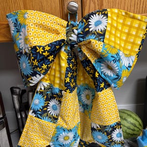 Puede incluir: Un gran lazo decorativo hecho de tela de patchwork con una mezcla de patrones en amarillo, azul marino y blanco. El diseño incluye cuadros vichy amarillos, pequeños estampados florales blancos y margaritas azules y blancas sobre fondo marino.