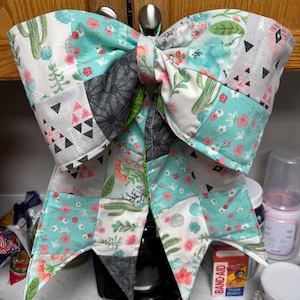 May include: A large fabric bow featuring a patchwork design with various patterns including green cacti, pink flowers, grey geometric triangles, and dark grey circular motifs on a white and teal background, with two long hanging tails at the bottom.