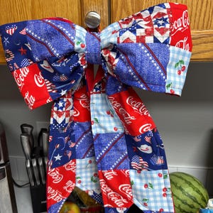 May include: A large fabric bow featuring a patchwork design with red, white, and blue colors. The pattern includes Coca-Cola logos, cherries, stars, and gingham checks. The bow has two long, pointed tails hanging down and is constructed from quilted fabric.