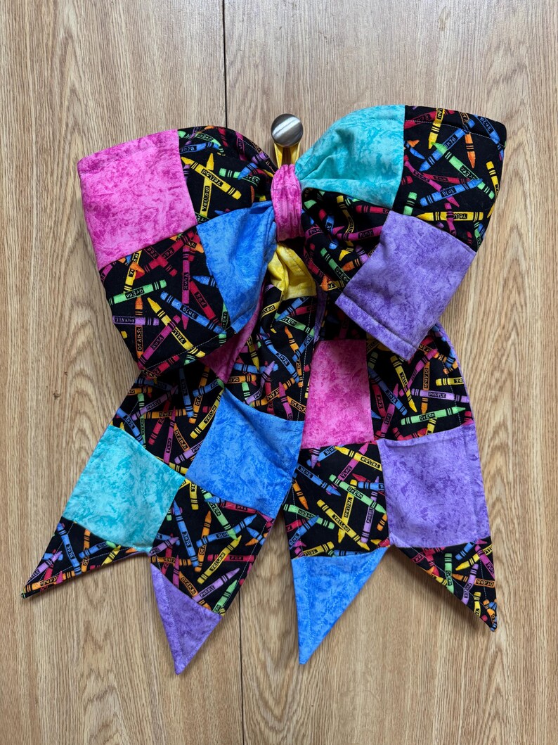 May include: A large fabric hair bow featuring a patchwork design of black fabric with a colorful crayon print, interspersed with solid textured squares in shades of pink, purple, and blue. The bow is secured with a central fabric knot.
