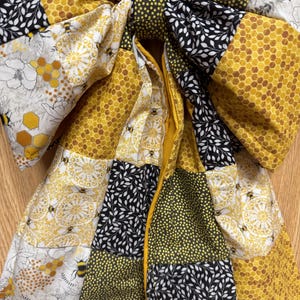 May include: A large fabric bow featuring a patchwork design of yellow honeycomb patterns, black and white leaf prints, yellow and black bumblebees, and small yellow polka dots on a dark background, with a central knot and two long hanging tails.