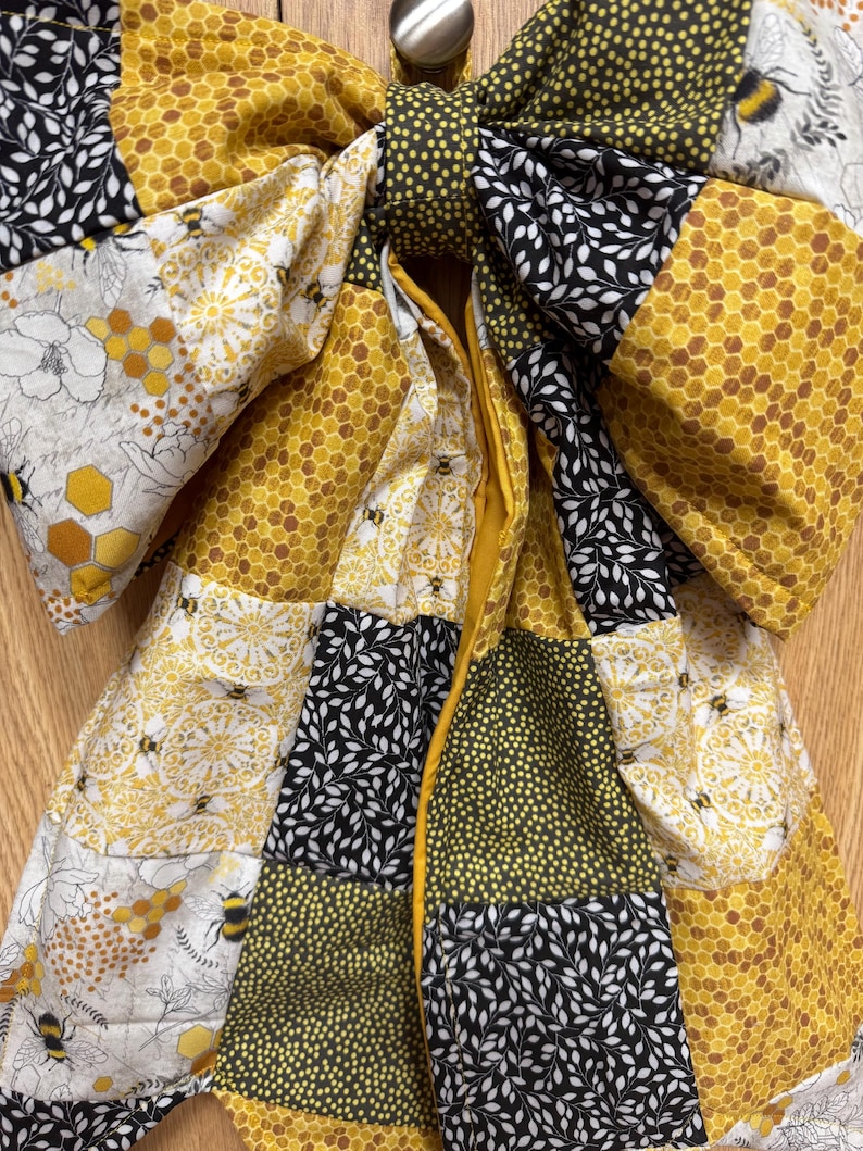 May include: A large fabric bow featuring a patchwork design of yellow honeycomb patterns, black and white leaf prints, yellow and black bumblebees, and small yellow polka dots on a dark background, with a central knot and two long hanging tails.