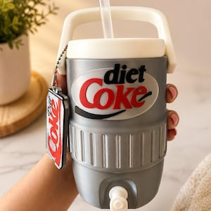 May include: A grey insulated beverage cooler with a white handle and lid, featuring the Diet Coke logo in red and black. A clear straw protrudes from the top, and a small Diet Coke branded charm hangs from a silver ball chain on the side.