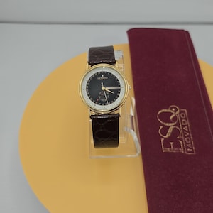 May include: A gold-tone wristwatch with a black circular dial, gold hands, and a dark brown textured leather strap. The watch face features the text ESQ MOVADO. Beside the watch lies a long, rectangular, burgundy velvet case with ESQ MOVADO in gold.