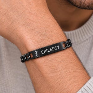 May include: A black metal medical alert bracelet featuring a curb chain design and a flat rectangular plate. The plate is engraved with a white medical caduceus symbol followed by the word EPILEPSY in clear, capitalized sans-serif lettering.