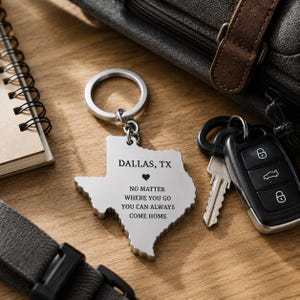 May include: A silver-toned metal keyring shaped like the state of Texas. The keyring features the engraved text DALLAS, TX, a small heart symbol, and the phrase NO MATTER WHERE YOU GO YOU CAN ALWAYS COME HOME in black lettering.