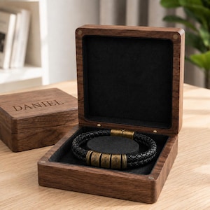 May include: A black braided leather bracelet with three antique brass cylindrical beads engraved with the names Carter, Daniel, and Alvaro, presented in a dark walnut wooden box with the name Daniel engraved on the lid.