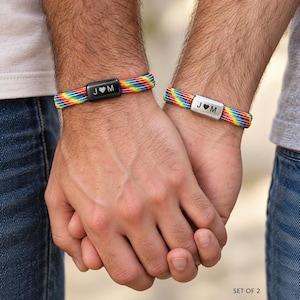 May include: A set of two bracelets featuring rainbow-colored woven cords. One bracelet has a black rectangular metal bead engraved with the text J heart M, and the other has a silver-toned rectangular metal bead engraved with the text J heart M.