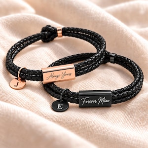 May include: Two braided black cord bracelets featuring metallic rectangular bars and circular charms. One bracelet has a rose gold bar engraved with Always Yours and a J charm. The other has a black bar engraved with Forever Mine and an E charm.