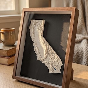 Op de afbeelding: Een driedimensionale reliëfkaart van Californië in wit, gemonteerd op een zwarte achtergrond in een houten schaduwdooslijst. De kaart toont gedetailleerde topografische texturen van het landschap, inclusief een klein blauw accentpunt.
