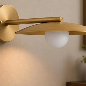 May include: A wall-mounted light fixture featuring a circular brass backplate, a horizontal arm, and a wide, flat, disc-shaped brass shade. A single spherical white glass bulb is positioned beneath the center of the shade, providing illumination.