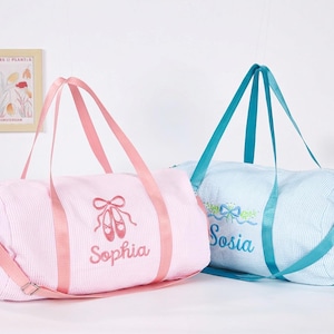 May include: Two cylindrical duffel bags with a pinstripe pattern. The left bag is light pink with pink straps and embroidered ballet slippers above the name Sophia. The right bag is light blue with blue straps and a floral bow above the name Sosia.